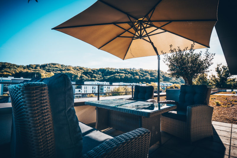 comitatus GmbH & Co.KG - EIne Terrasse mit Sonnenschirm, Tisch und Stühlen vor einer grünen Landschaft bei Sonnenschein